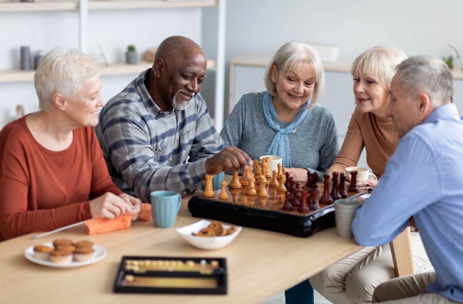 A group of older adults sitting next to each other at a table playing a game of chess in a senior living community.