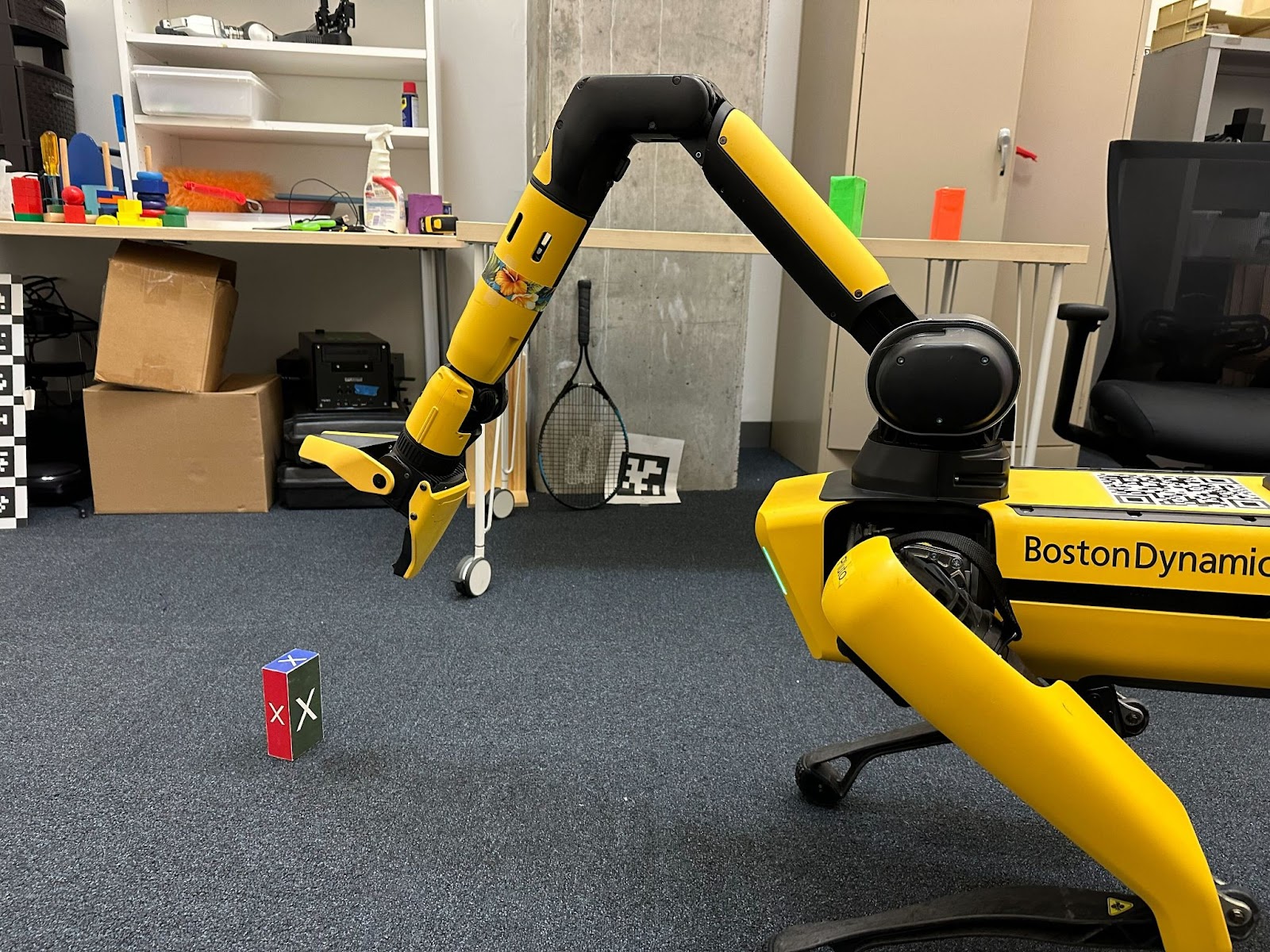 A yellow-and-black Robot reading "Boston Dynamics" on one side poses in the foreground of the photo, the long arm attached to its "head" reaching forwards to pick up a small rectangular box with colorful sides emblazoned with the letter X.
