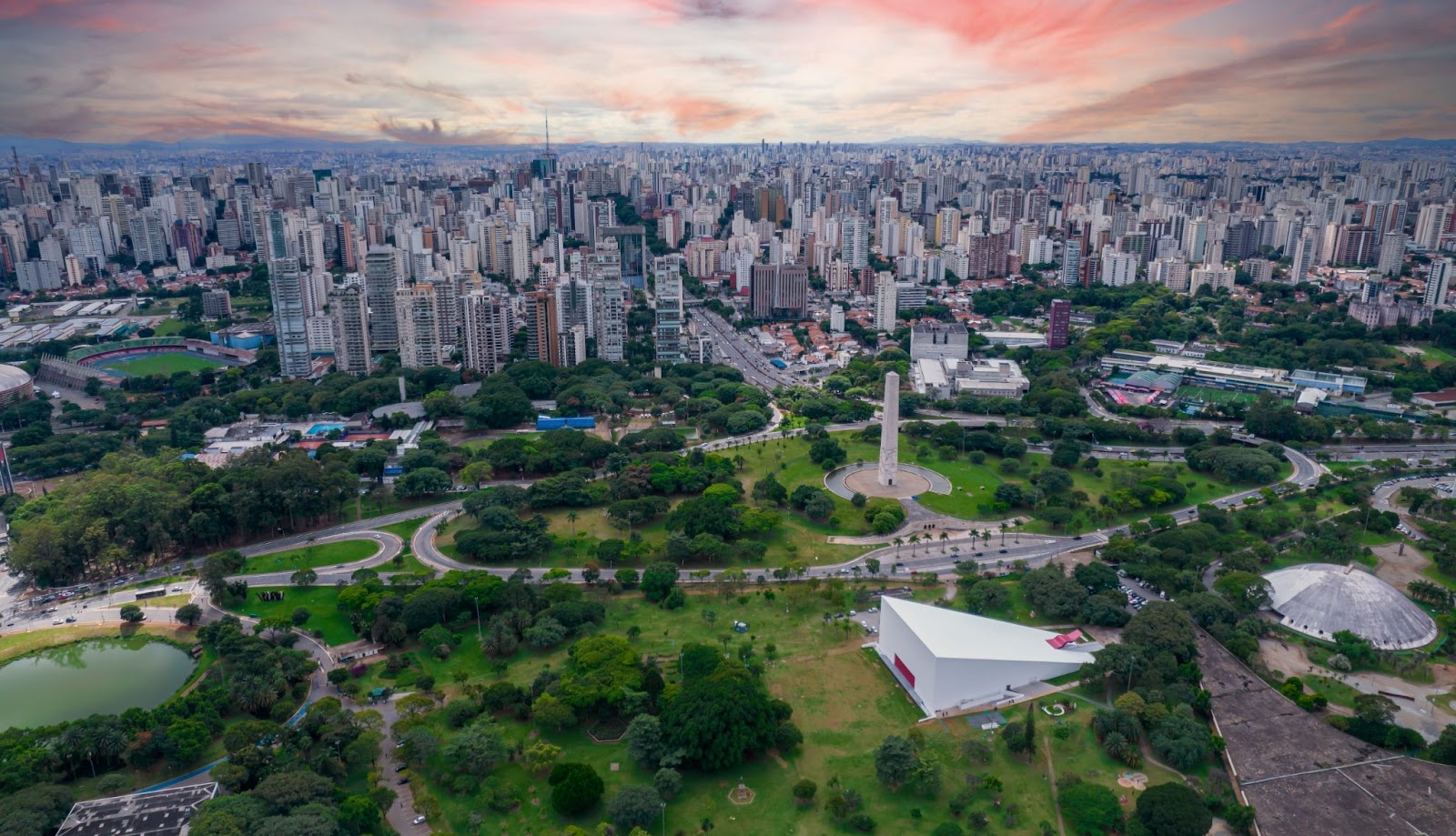 Imagem aérea do Parque Ibirapuera. Vasta área verde com lago, árvores e gramado. Ao horizonte, diversos prédios de diferentes tamanhos.