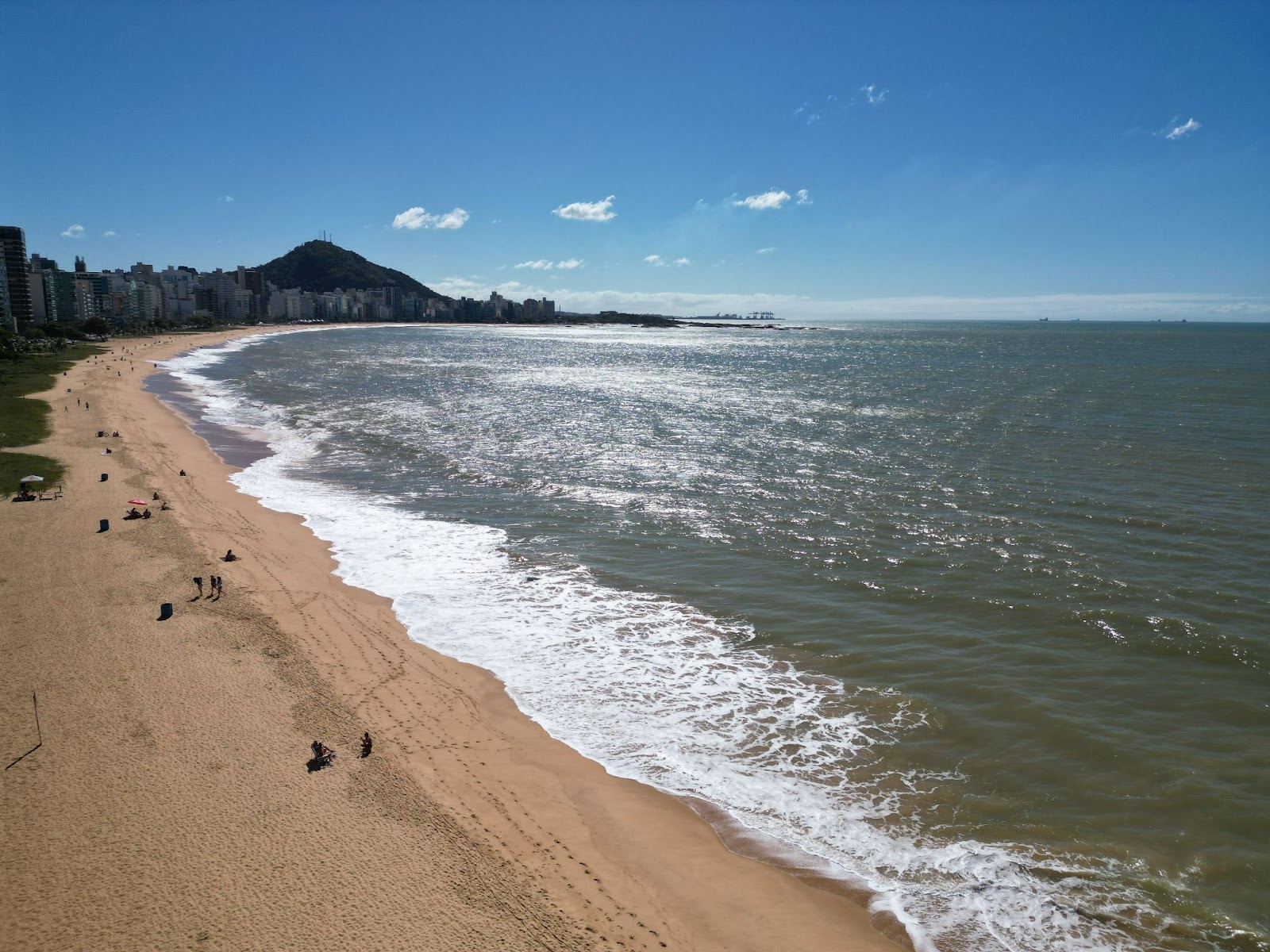 Vista aérea da Praia da Costa, com o mar ao lado direito da imagem em tom esverdeado, diferente do céu azul, chegando na extensa faixa de areia.
