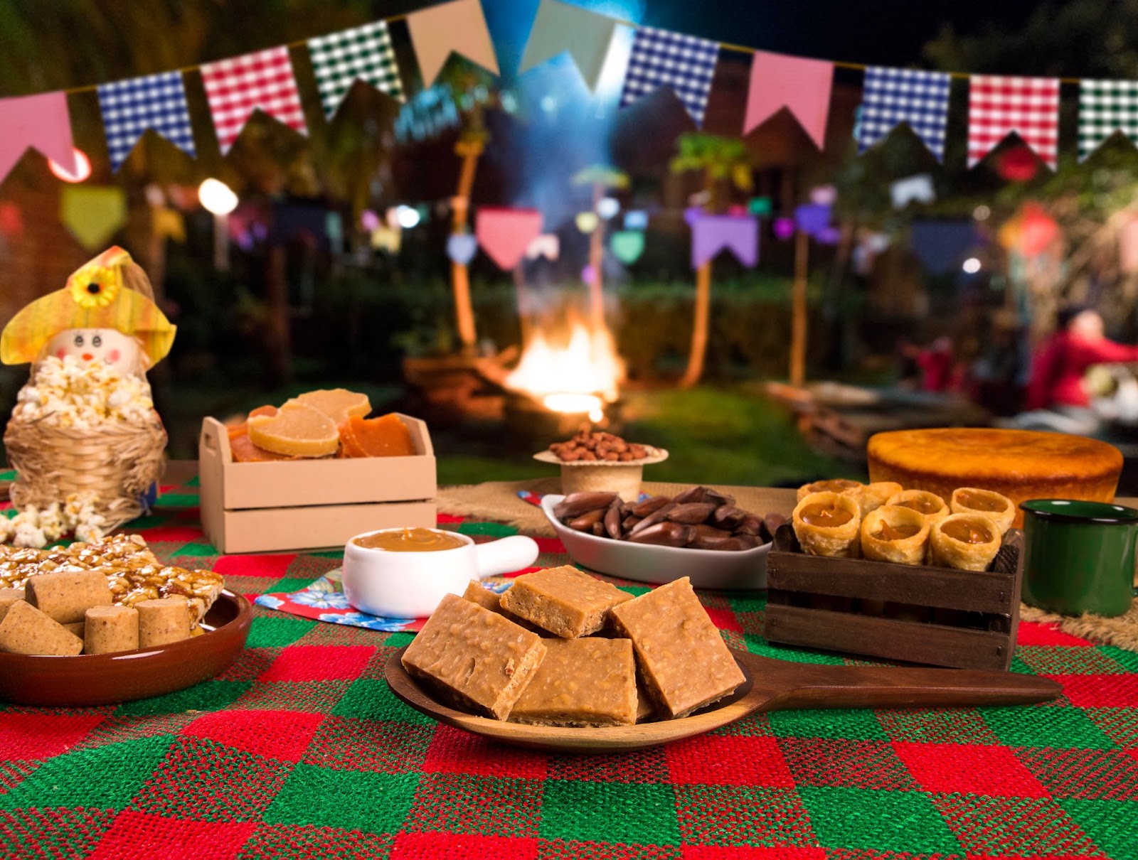Mesa com toalha quadriculada verde e vermelha. Sobre ela estão vários pratos típicos juninos como pinhão, paçoca e bolo de milho. Ao fundo, aparece uma fogueira desfocada e algumas bandeirolas coloridas.
