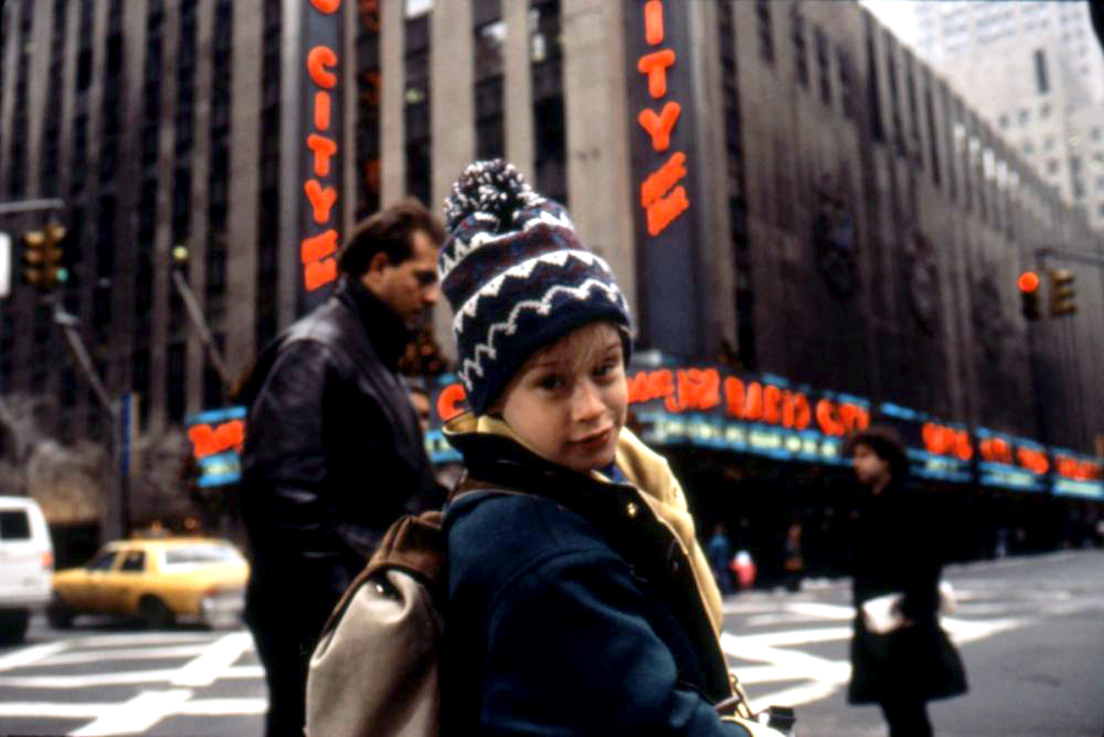 Kevin McCallister, portrayed by Macaulay Culkin, turns back to the camera with a mischievous grin outside Radio City Music Hall in Home Alone 2, evoking the playful and heartwarming moments reminiscent of the best comfort Christmas films. His expression hints at a clever plan or a playful moment amidst the bustling cityscape of New York, adding to the film's festive and charming ambiance.