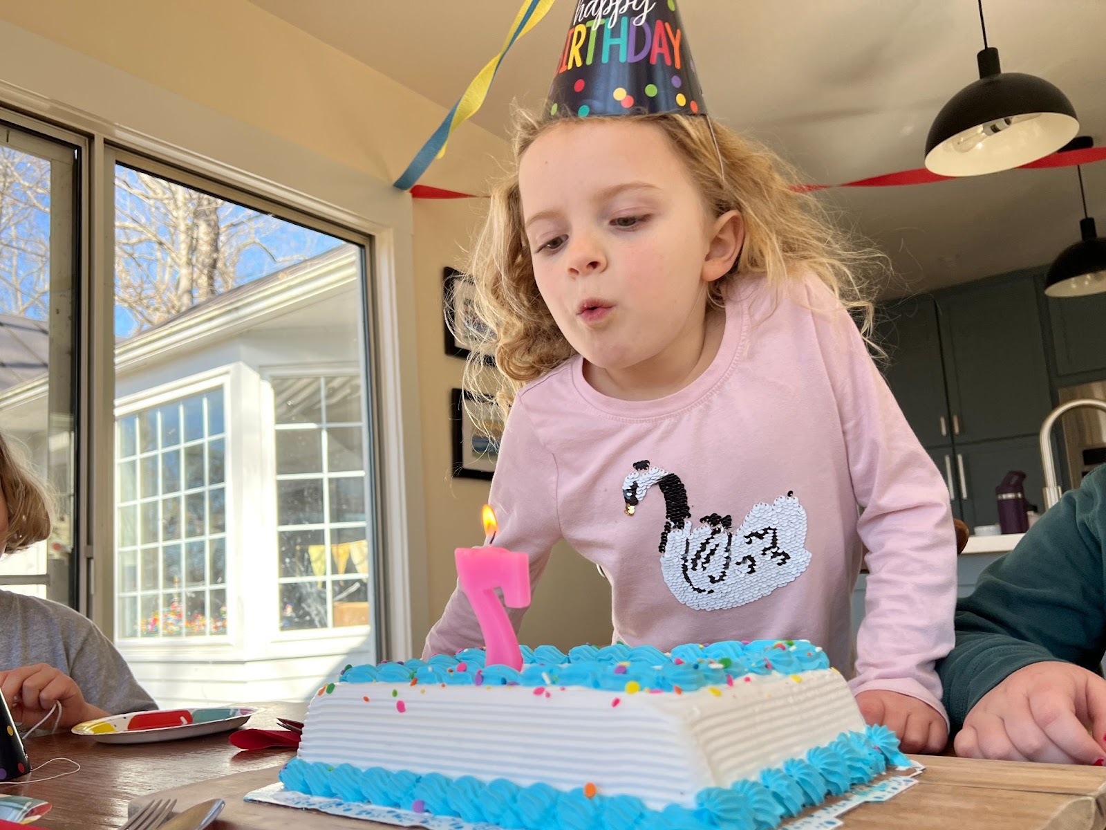 A little girl in a pink shirt with a swan on the front blows out a candle on a birthday cake shaped like the number 7.
