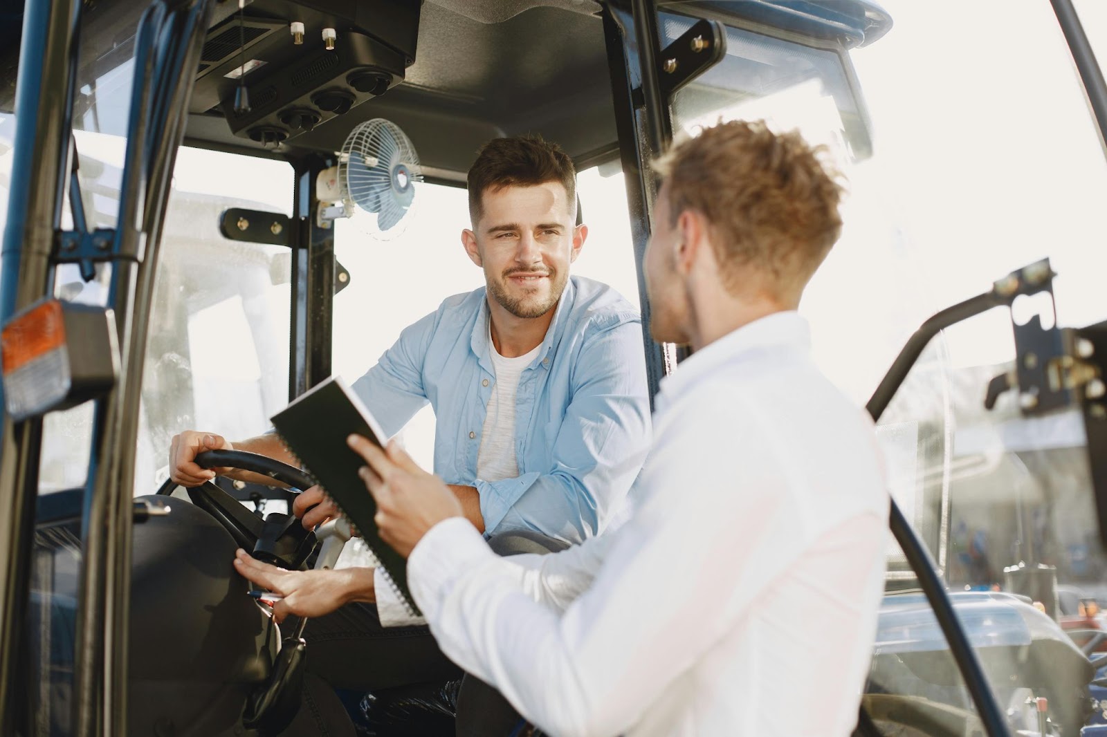 Two people talking to each other — one sitting behind the wheel of a forklift and the other pointing at a notebook