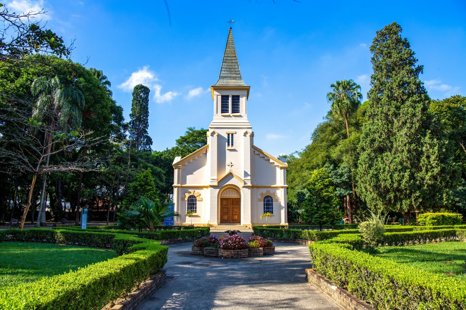 Pequena capela branca com detalhes em amarelo e porta de madeira em estilo colonial, em frente ao jardim do Parque Vicentina Aranha.