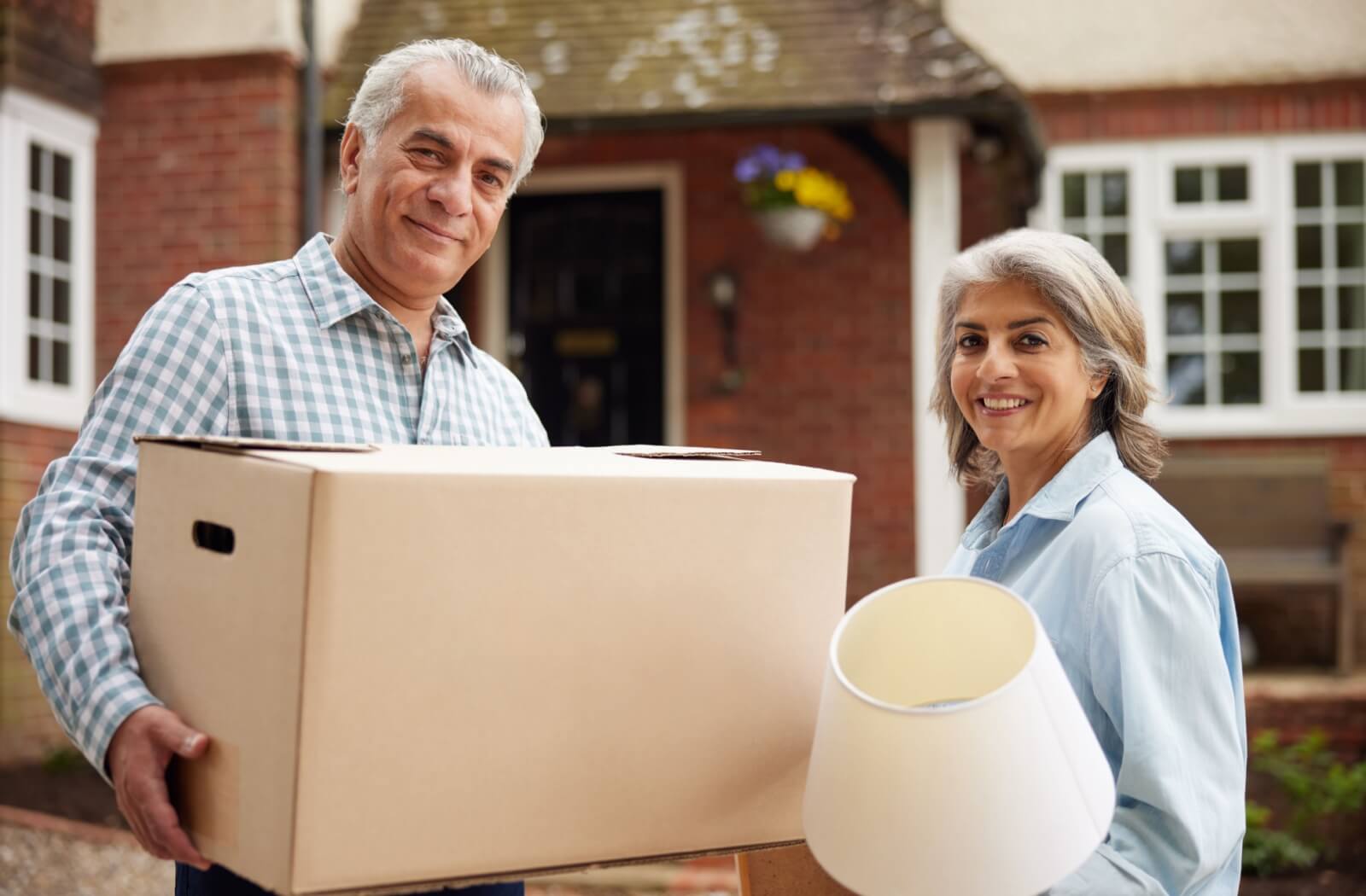 An older adult couple carrying a box and a lamp as a part of their downsizing efforts.