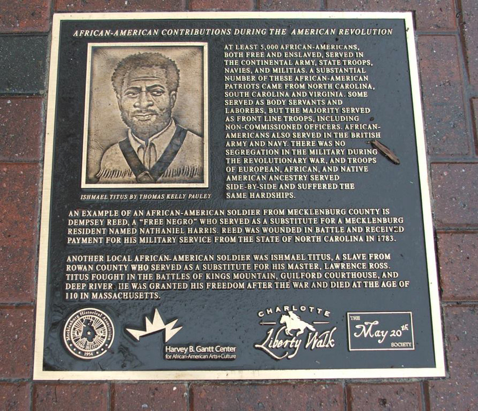 A plaque on a sidewalk. It depicts Ishmael Titus: he is middle aged with medium, bushy hair. He has a beard. He is wearing a buttoned shirt with a deerskin jacket.