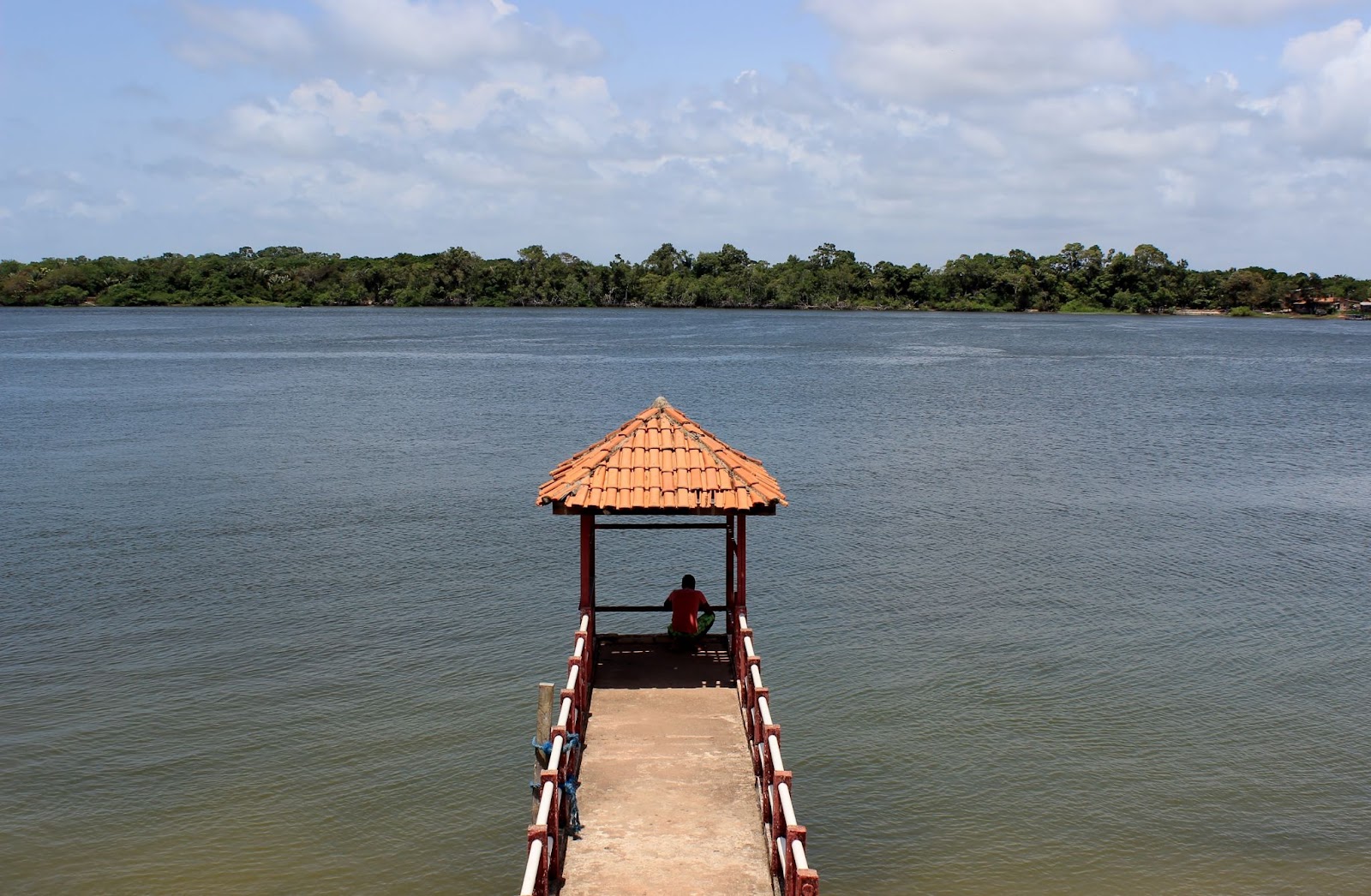 Píer na Baía de Marajó, Ilha de Marajó. O estreito caminho de cimento avança sobre a água escura do rio e termina em um pequeno gazebo de telha laranja. Ao fundo, aparecem as matas verdes.