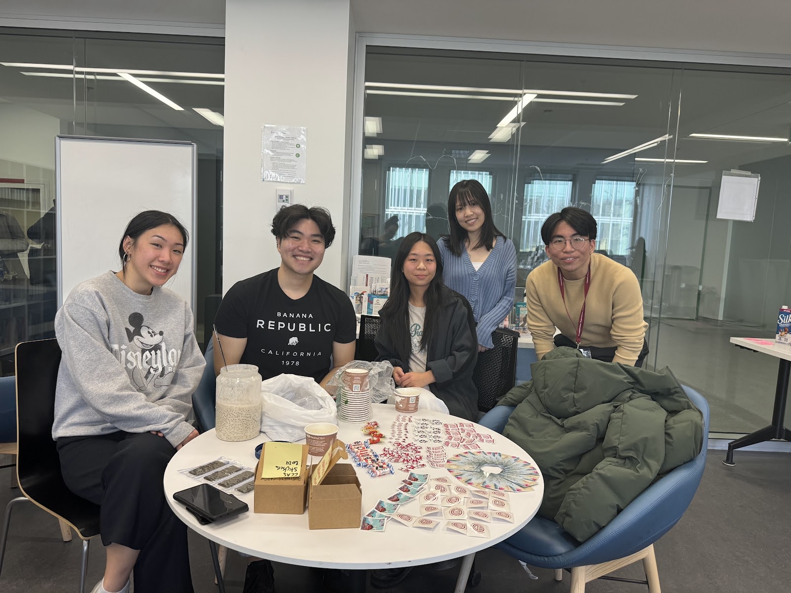 Five Asian students sit and stand around a white table. A large container of sago dessert soup (which is white, with small clear coloured pearls) is on the table along with many colourful stickers and candies.