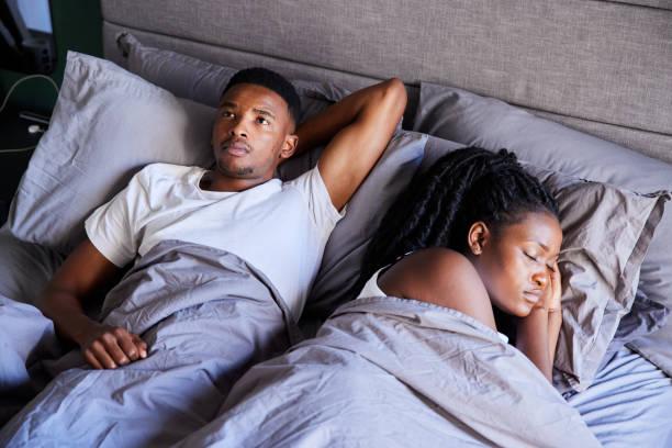 young man looking unhappy while lying in bed with his sleeping wife - black sad man stock pictures, royalty-free photos & images