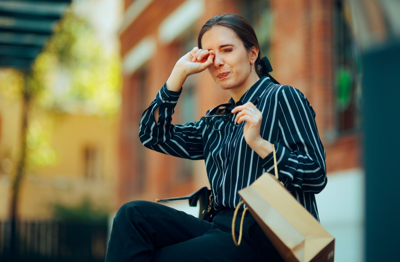 A woman sitting on a bench and rubbing her itchy eye with her right hand