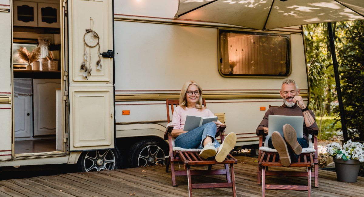 Senior couple enjoying leisure time while relaxing on wooden chairs outside their camper van, with the man using a laptop and the woman reading a book, surrounded by lush greenery.