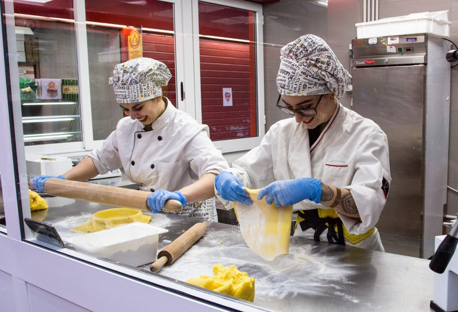 A couple of women in chef's coats making dough

Description automatically generated