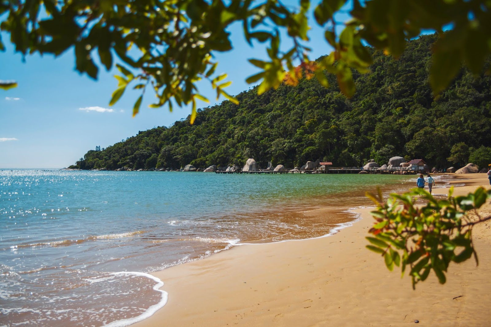 Praia das Laranjeiras. O mar tranquilo tem uma tonalidade turquesa e a faixa de areia está quase vazia, com exceção de duas pessoas que caminham por ela. A orla acaba em um morro coberto por mata verde