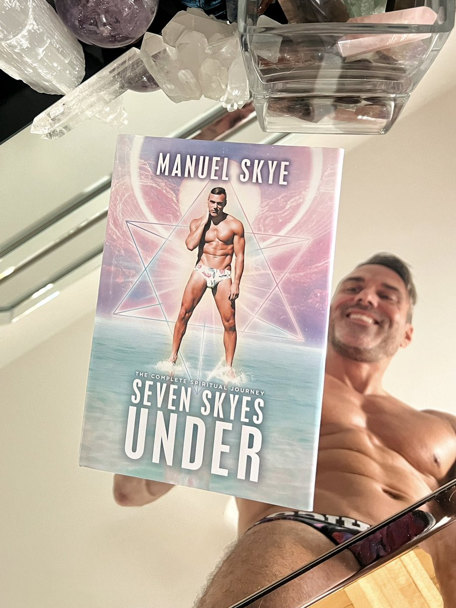 Manuel Skye posing and smiling over a glass coffee table with his spiritual self help book seven skyes under pressed cover down on the table in the foreground of the photo