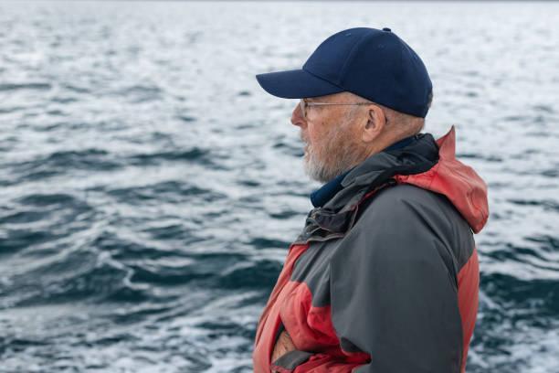 Ahoy Mate! A senior man enjoying a tourist excursion while on holiday in Torridon, Scotland. He is exploring the seas on a tourboat and looking at the view with a contented look on his face. baseball cap stock pictures, royalty-free photos & images