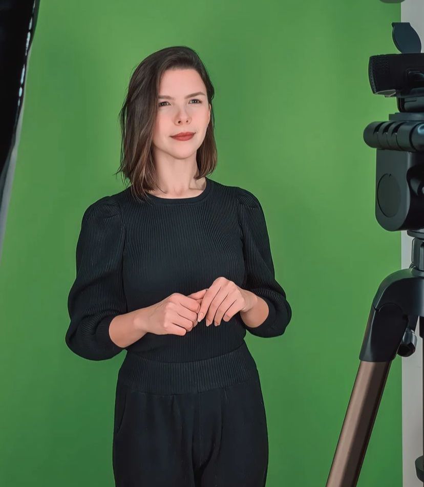 foto de Hanna, tradutora e intérprete de Libras -português posicionada em estúdio profissional de captação de imagem. Hanna é mulher branca, de cabelos castanhos curtos e veste blusa e calça preta, Sorri suavemente para uma câmera sobre tripé à sua frente.
