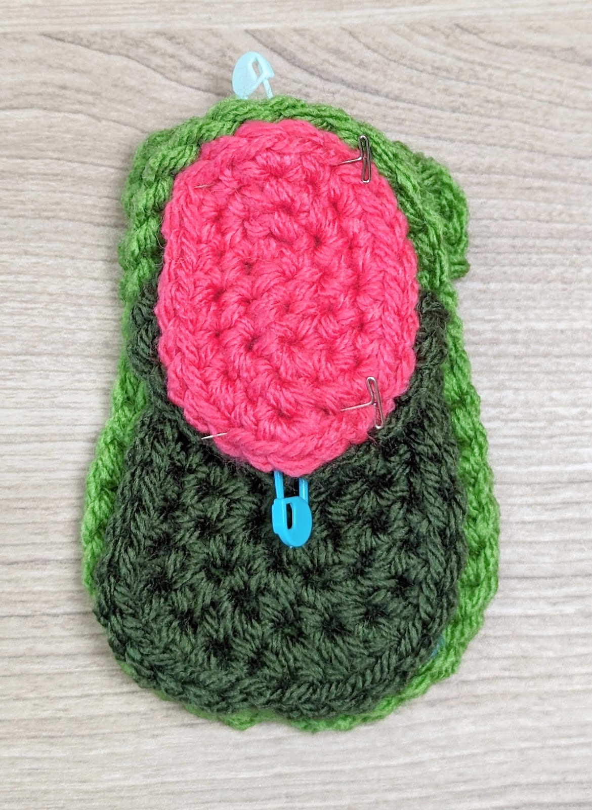 A crochet avocado made with green and dark green yarn for the body and pink yarn for the seed, secured with blue and silver stitch markers, lies on a light wooden surface.