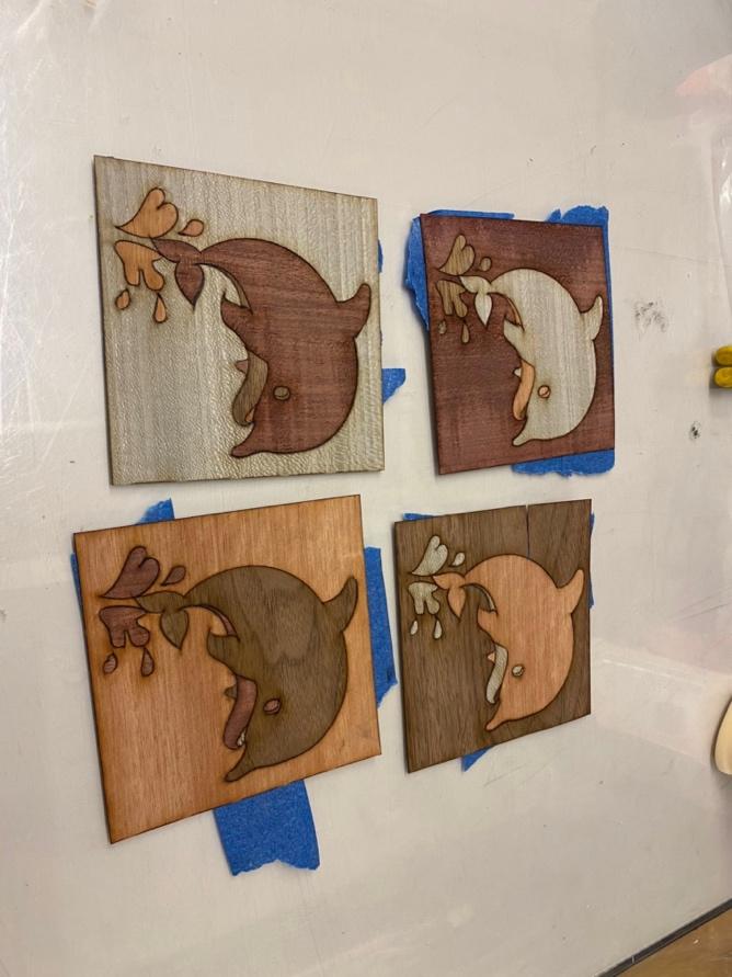 Four wooden blocks are taped to a table with painter's tape. Each one is etched with a leaping dolphin, made from different shades and textures of wood.