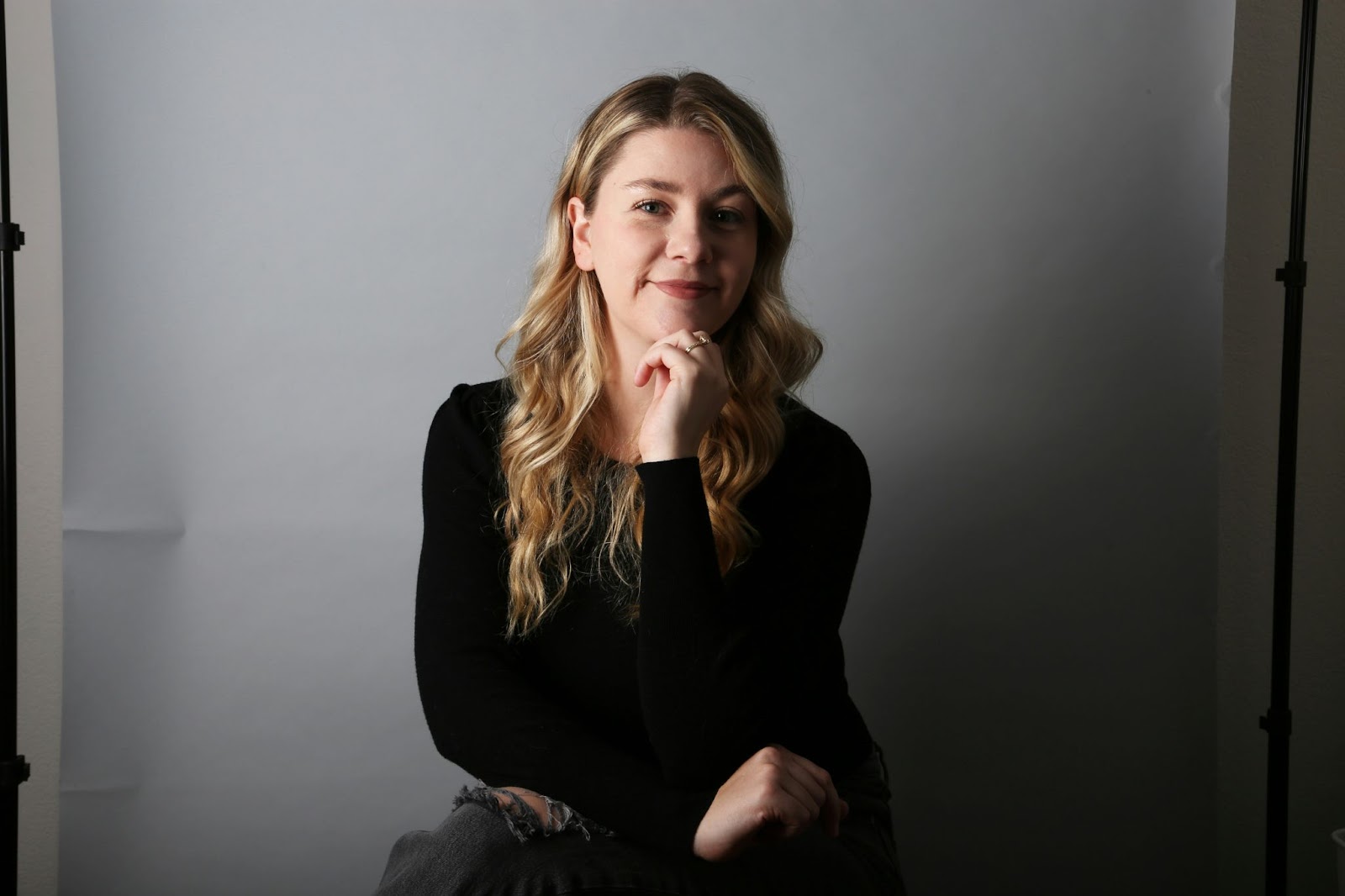 A woman giving the hand-on-chin pose is sitting in front of a white paper background to get clicked