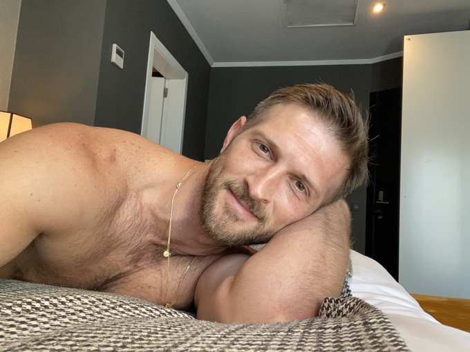 gay muscle daddy Paul Wagner posing naked on the bed and smiling for the camera in an iphone selfie