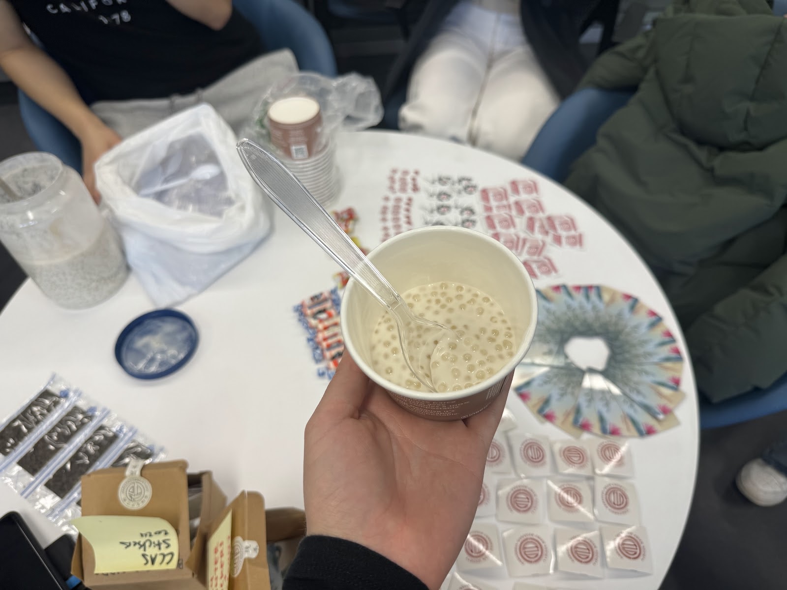 A white person’s hand holds a compostable cup filled with a white soup, which contains clear sago pearls. Some stickers are visible on the table in the background.