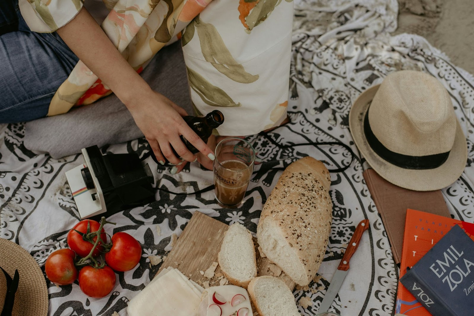 picnic blanket with food and bottled beverage along with books camera and hat