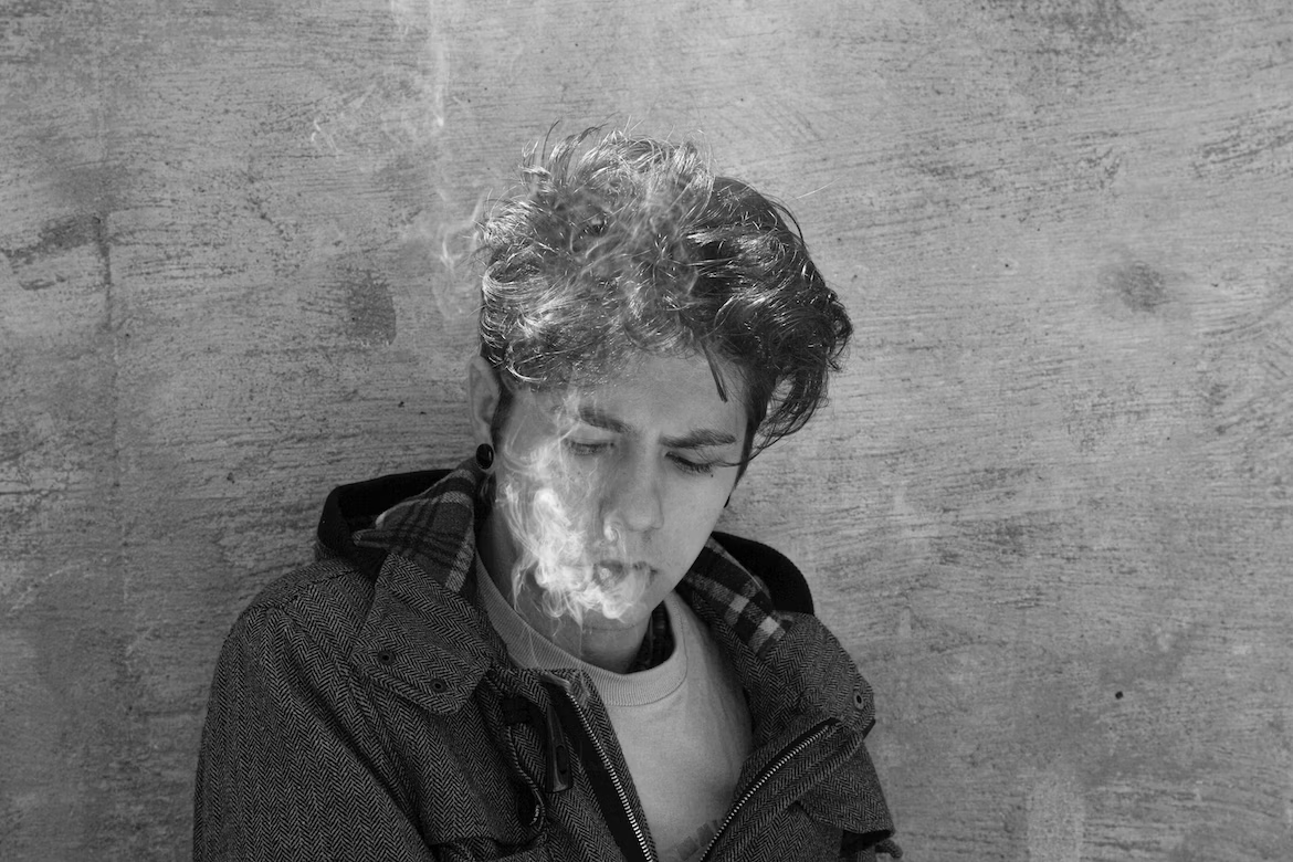 a man in a jacket leaning against a wall smoking a cannabis joint