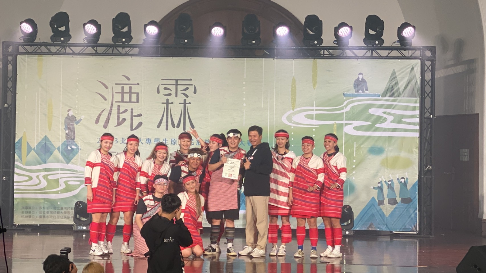 一群人穿著原住民服飾在舞台上表演完合照，背景板寫著「漉霖」，大家笑得很開心。