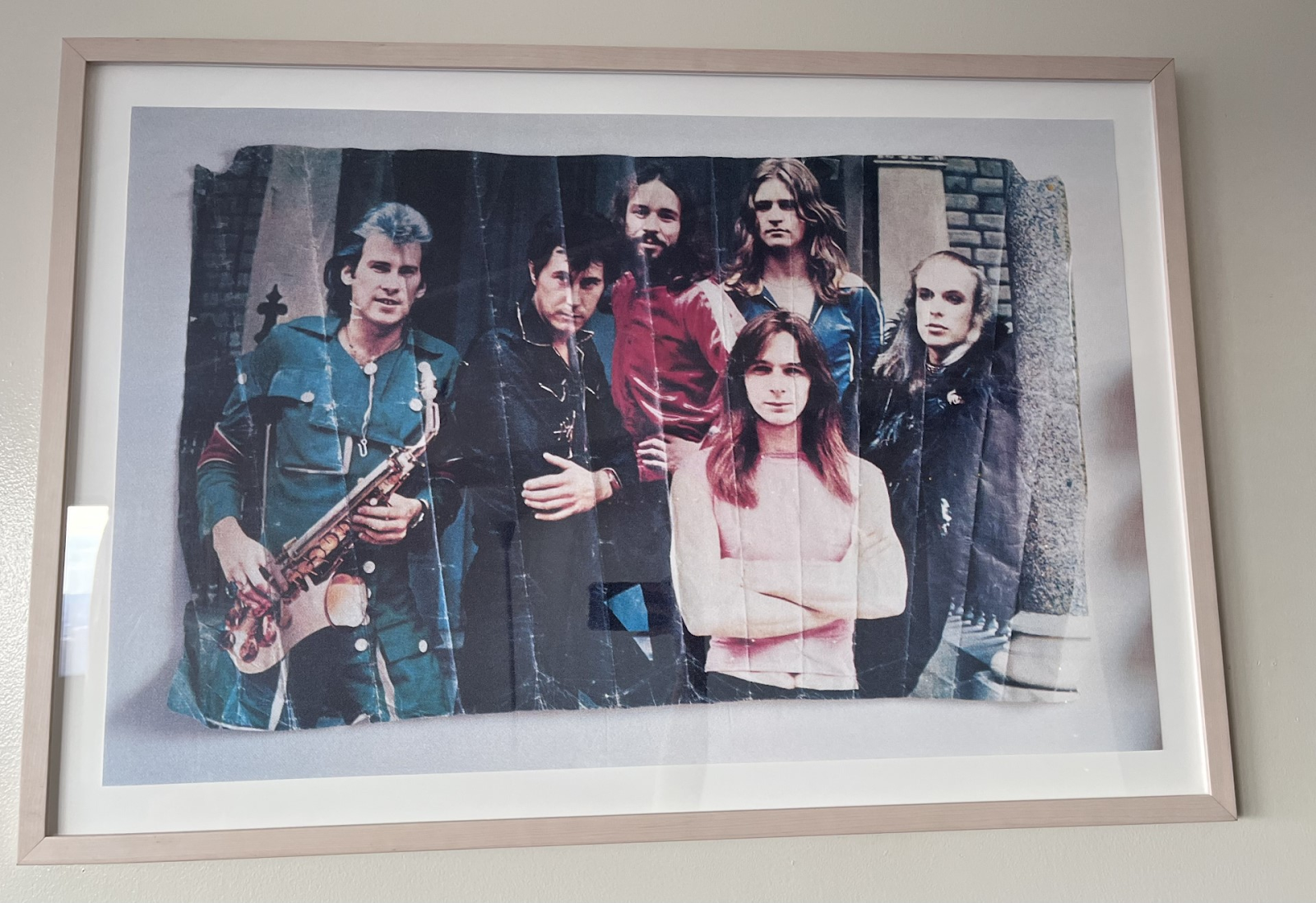 The framed art piece, which features a photo print of a vintage photo print that has been crinkled and folded, but is now stretched out for the viewer to see. Six band members pose, facing the camera, some smiling and others staring down the photographer.