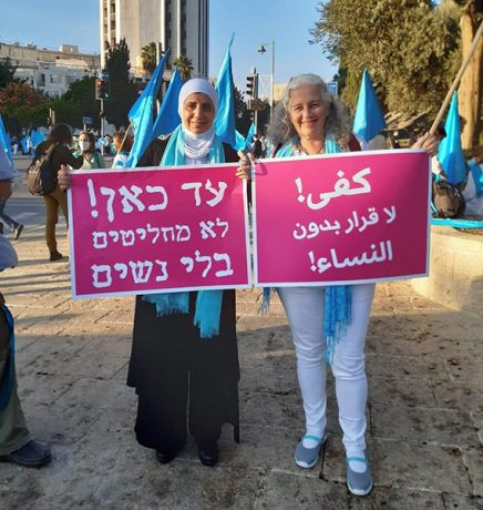 A woman in hijab holds up a sign in Hebrew, a woman without hijab holds up a sign in Arabic.
