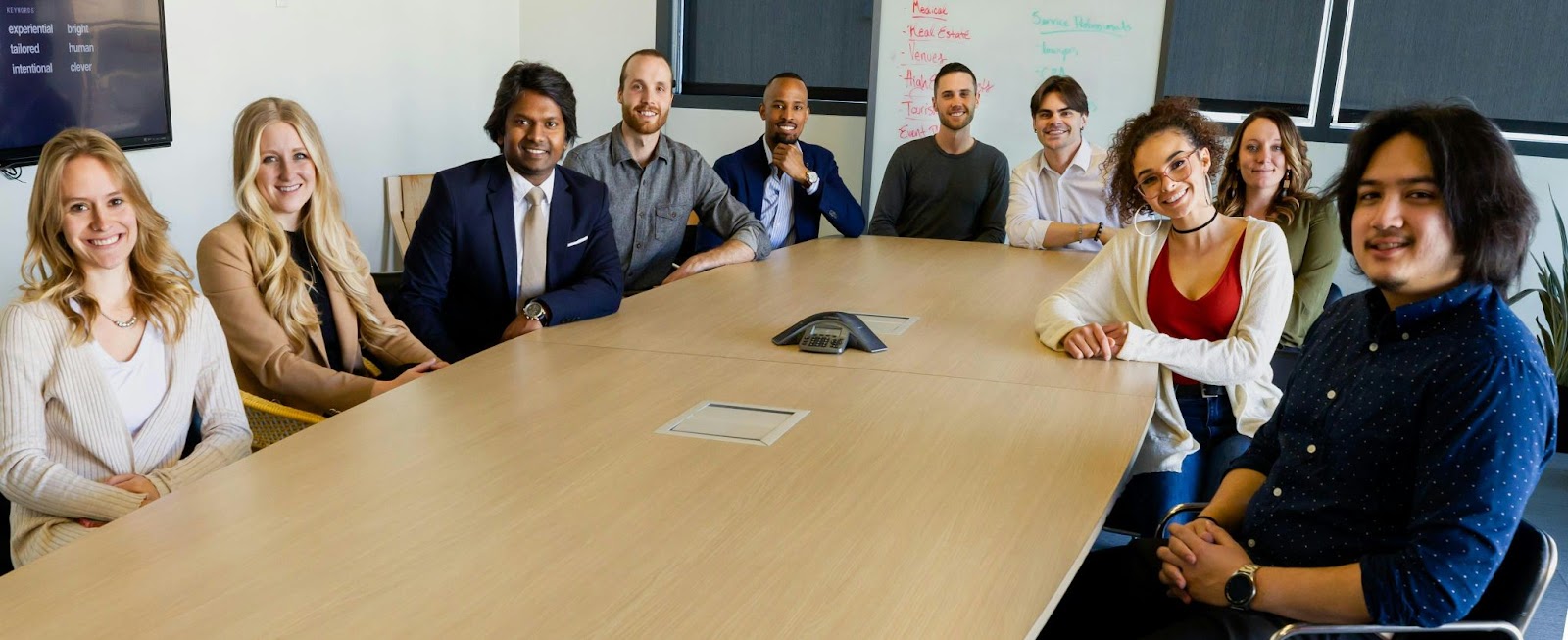 A group of employees of different races sited at a round table. They are all smiling. They were likely taking a company group photo. (Diversity tips blog)