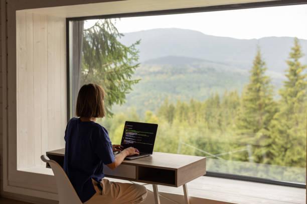 Woman works on laptop remotely in house on nature Woman works on laptop while sitting by the table in front of panoramic window with great view on mountains. Remote work and escaping to nature concept remote access stock pictures, royalty-free photos & images