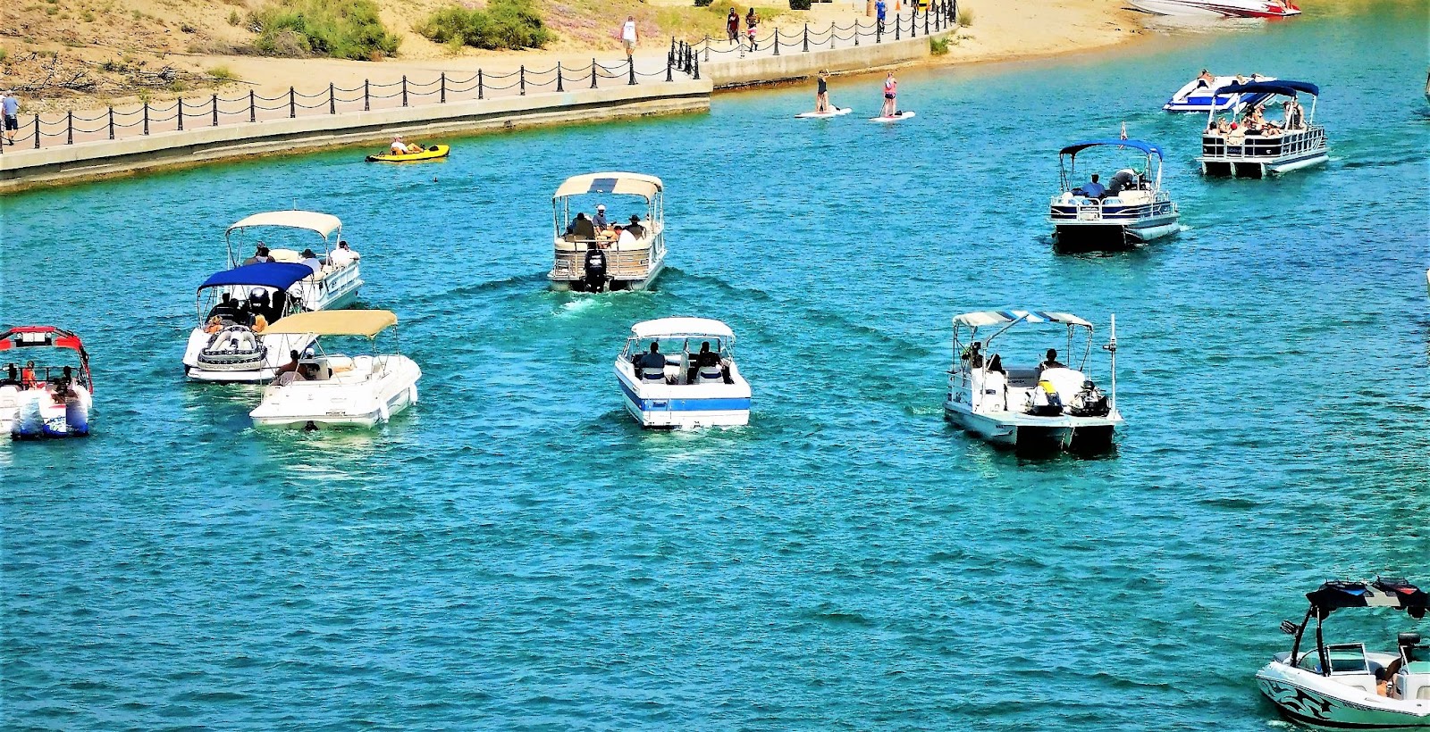 Several pontoon boats float on a lake. In the background, the shoreline is visible and people walk along a path.
