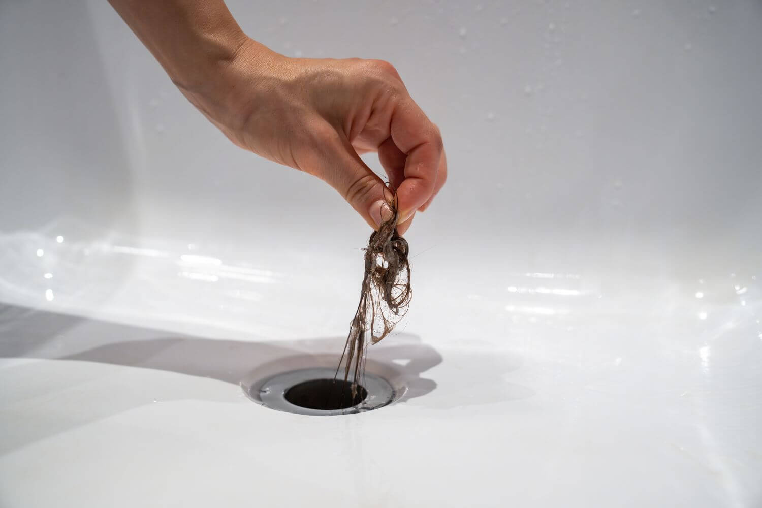 Hand pulling up hair from a shower drain in a white bathtub