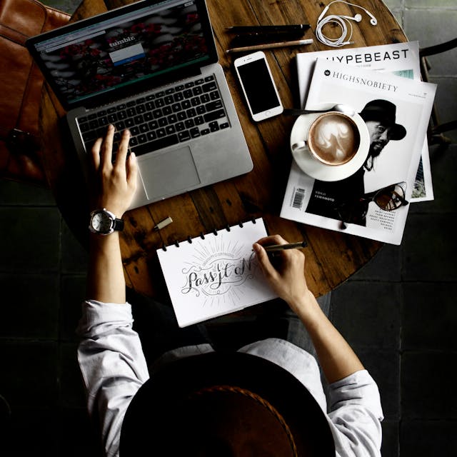 A top view of a man working on a font design for UI UX in a notepad in front of a laptop with magazines and coffee beside him