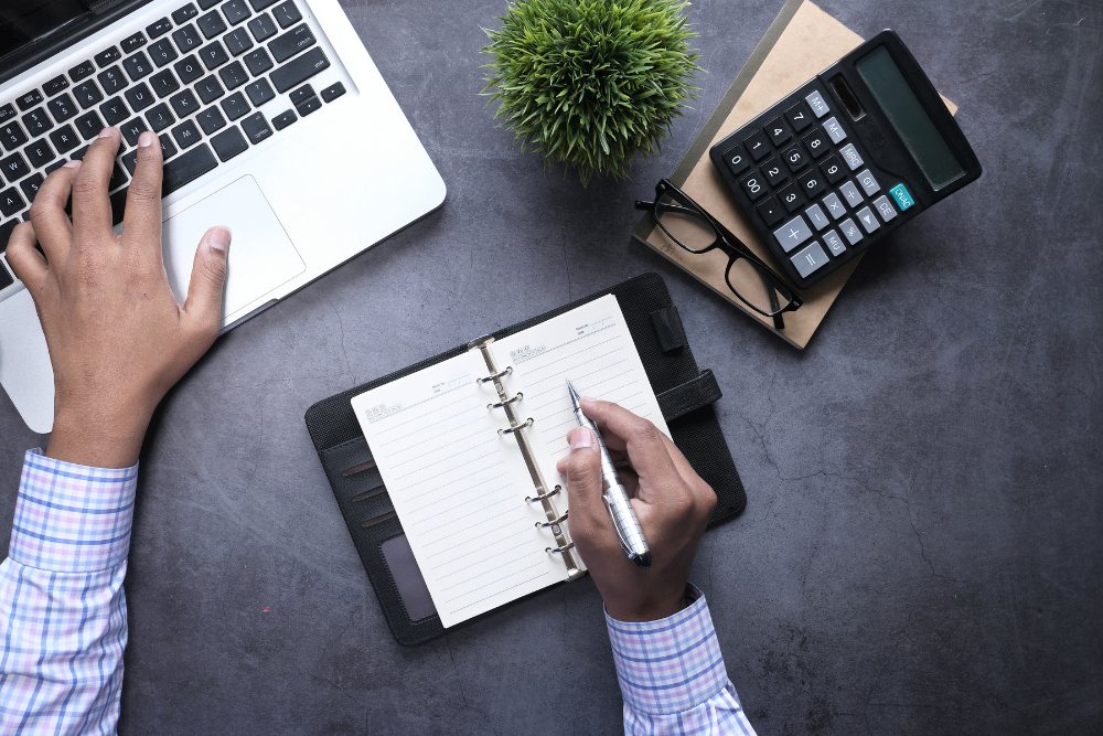 Foto de uma pessoa com um notebook e uma agenda calculando o valor de entrada de um apartamento sobre uma mesa de cor cinza escuro, também tem uma calculadora e óculos em cima da mesa.