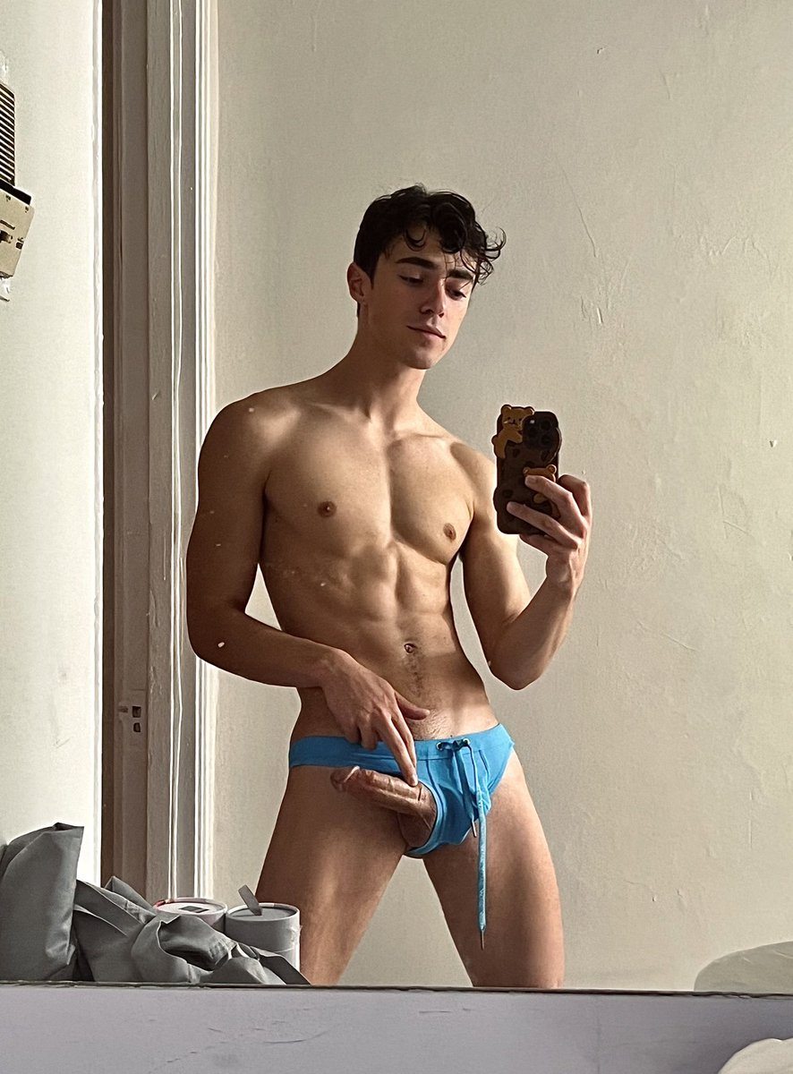 Nick Floyd taking a shirtless mirror selfie in a blue speedo with his erect dick peeking out of his swimsuit