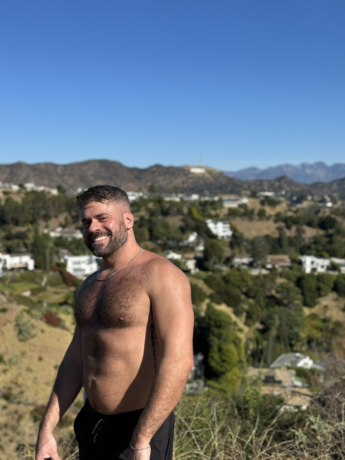 Max Romano standing shirtless outside smiling for a photo while wearing black sweat shorts