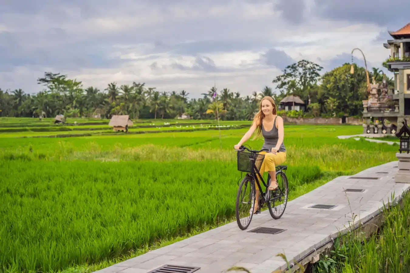 A smiling woman on a bicycle rides along a path next to vibrant green rice terraces. The backdrop features traditional architecture, suggesting a serene, rural setting, likely in a Southeast Asian countryside.