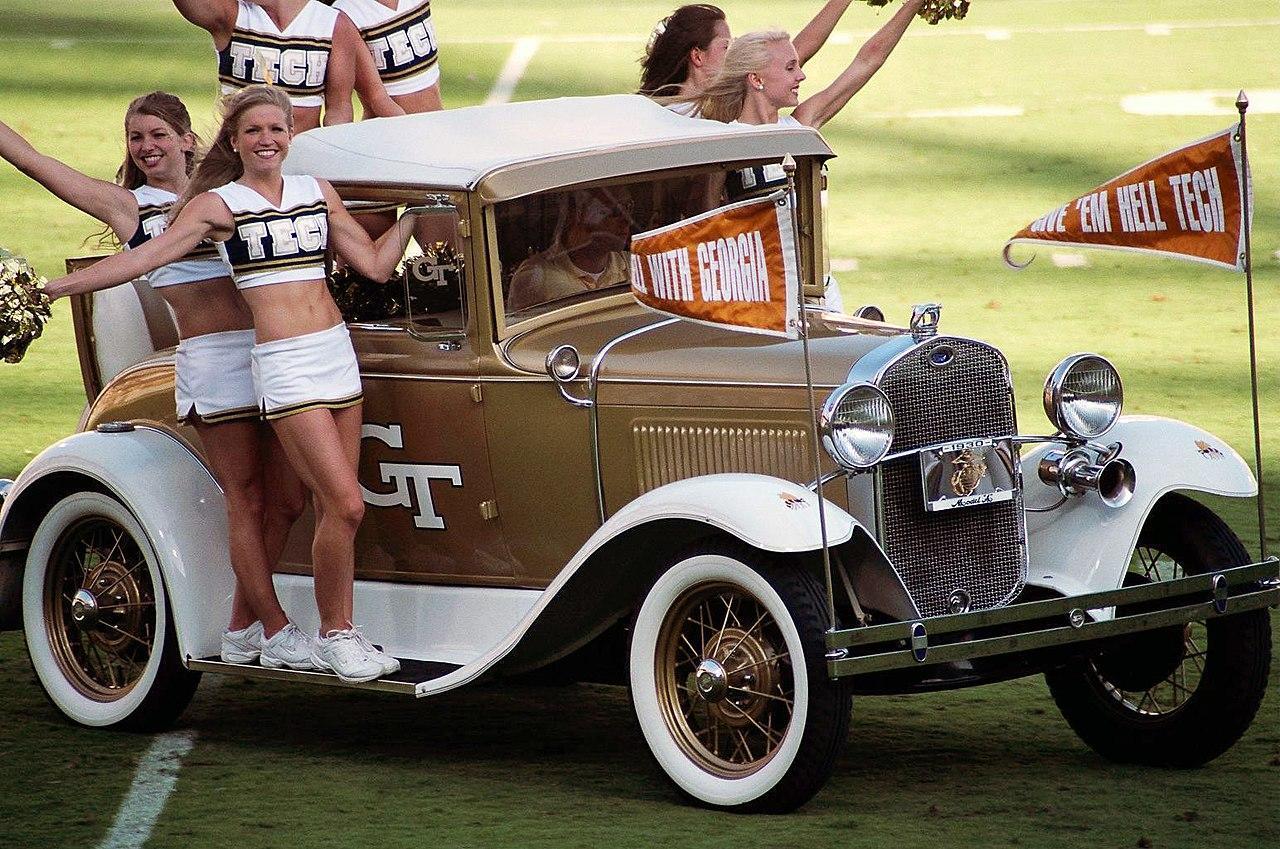 Six women, wearing a uniform of a white skirt and a white and gold cropped top with the word "Tech" on the front, ride onto the football field on the running boards and rear seat of a white-and-gold-painted antique car.