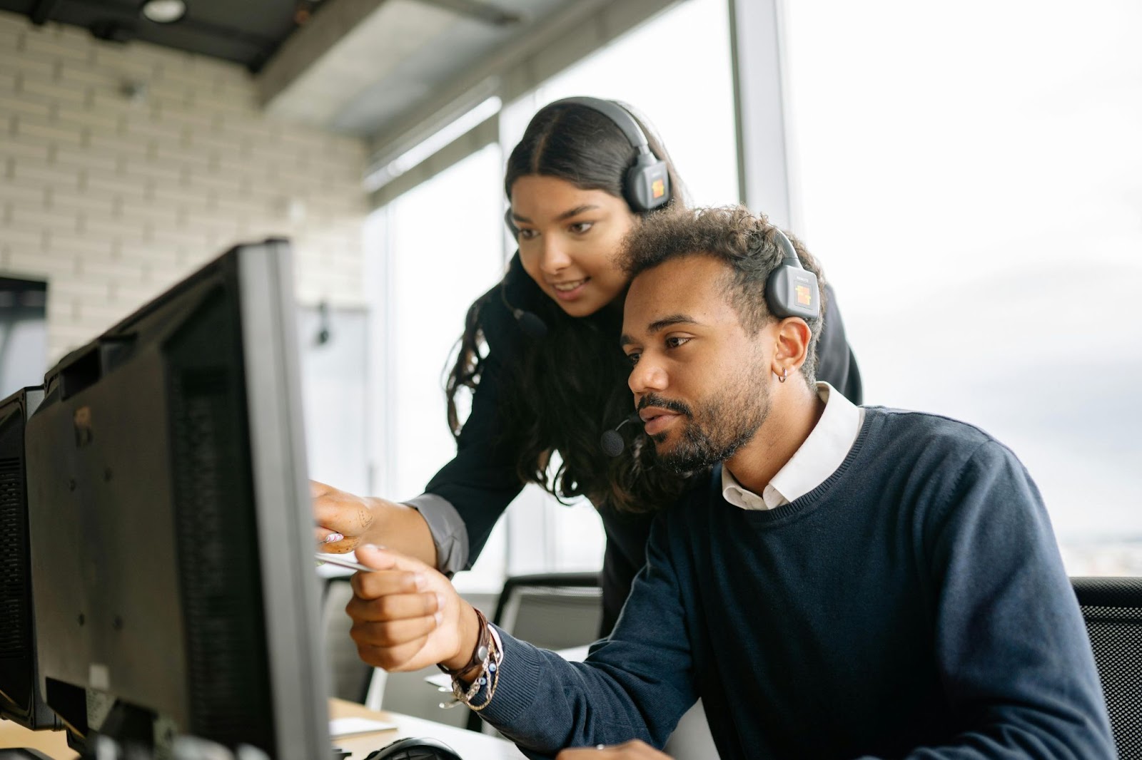 A man sitting on his desk chair and a woman looking over him, both of them wearing headphones and pointing at the computer screen.