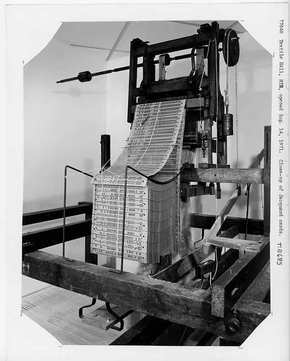Image: [Fig.4.] A black and white archival photo of the Jacquard Loom, a tall device with long wooden slats stacked against each other, running up through an opening near the top. Photo courtesy of the National Museum of American History.