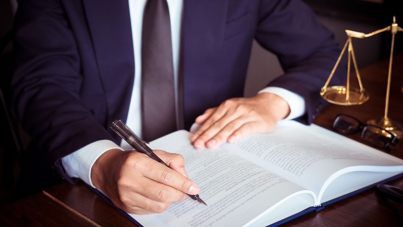 Close-up image of a person's hands, one holding a pen, poised to write in a large open book on a wooden table. The individual appears to be wearing a formal dark suit and a white shirt with cufflinks. In the background, a scale of justice is in focus, symbolizing legal practice. The presence of glasses next to the book suggests a studious or professional setting, possibly in a law office.