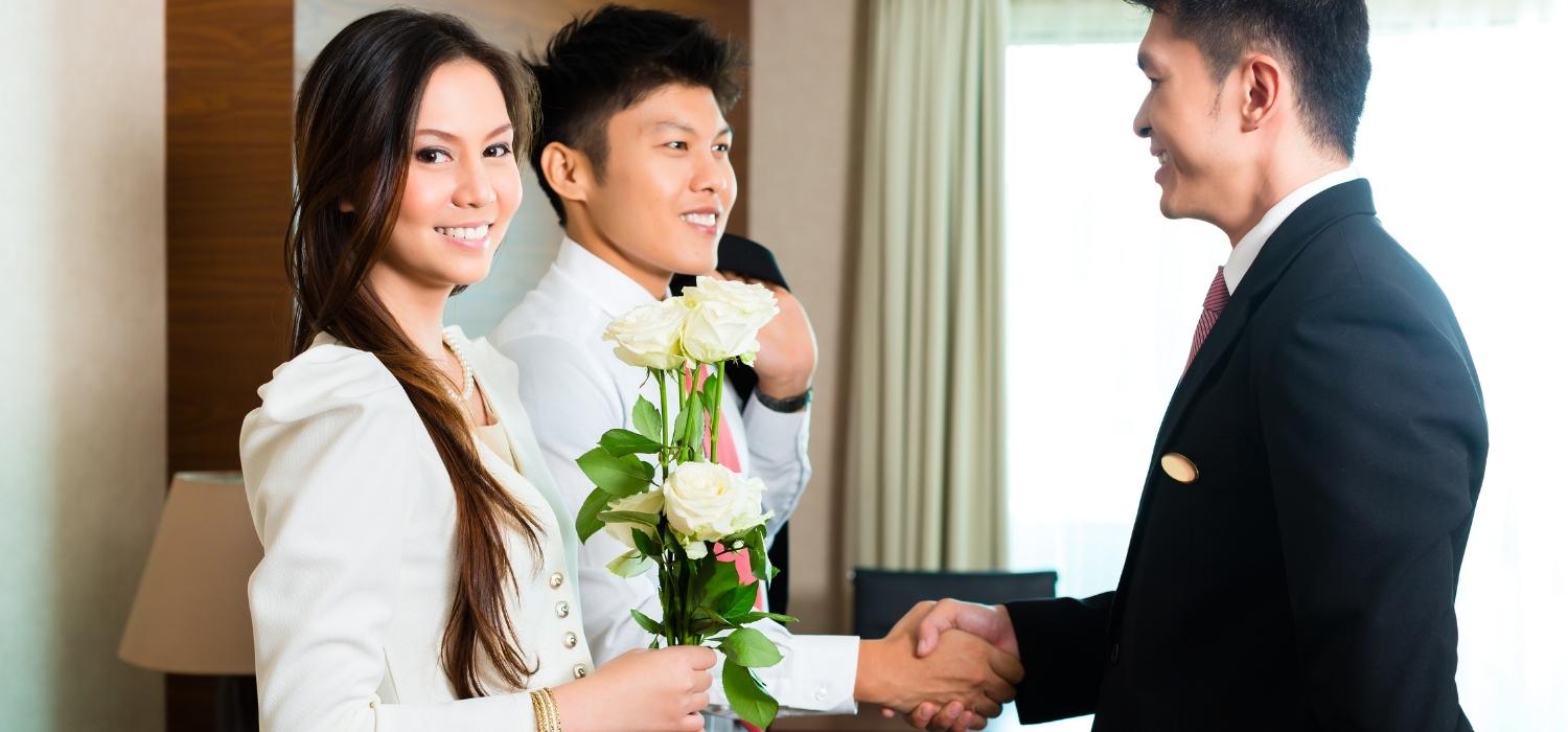 A smiling woman is facing the camera, carrying white roses, and her fiancé is booking a hotel room meeting with the owner.