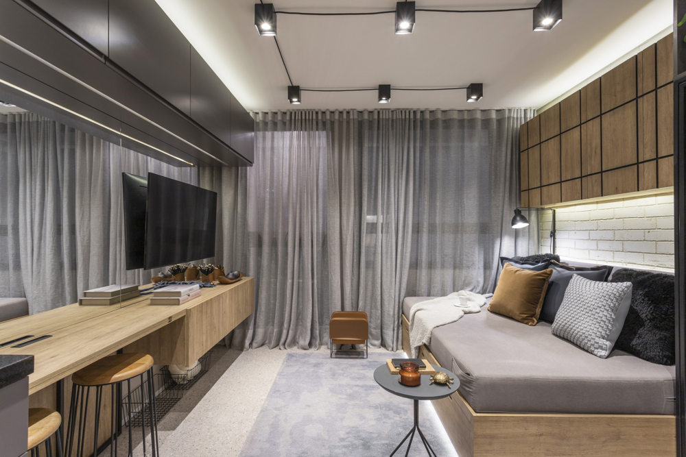 Sala com decoração industrial, com cortina e tapete na cor cinza, com uma bancada de madeira fixada na parede com banquetas e uma televisão.