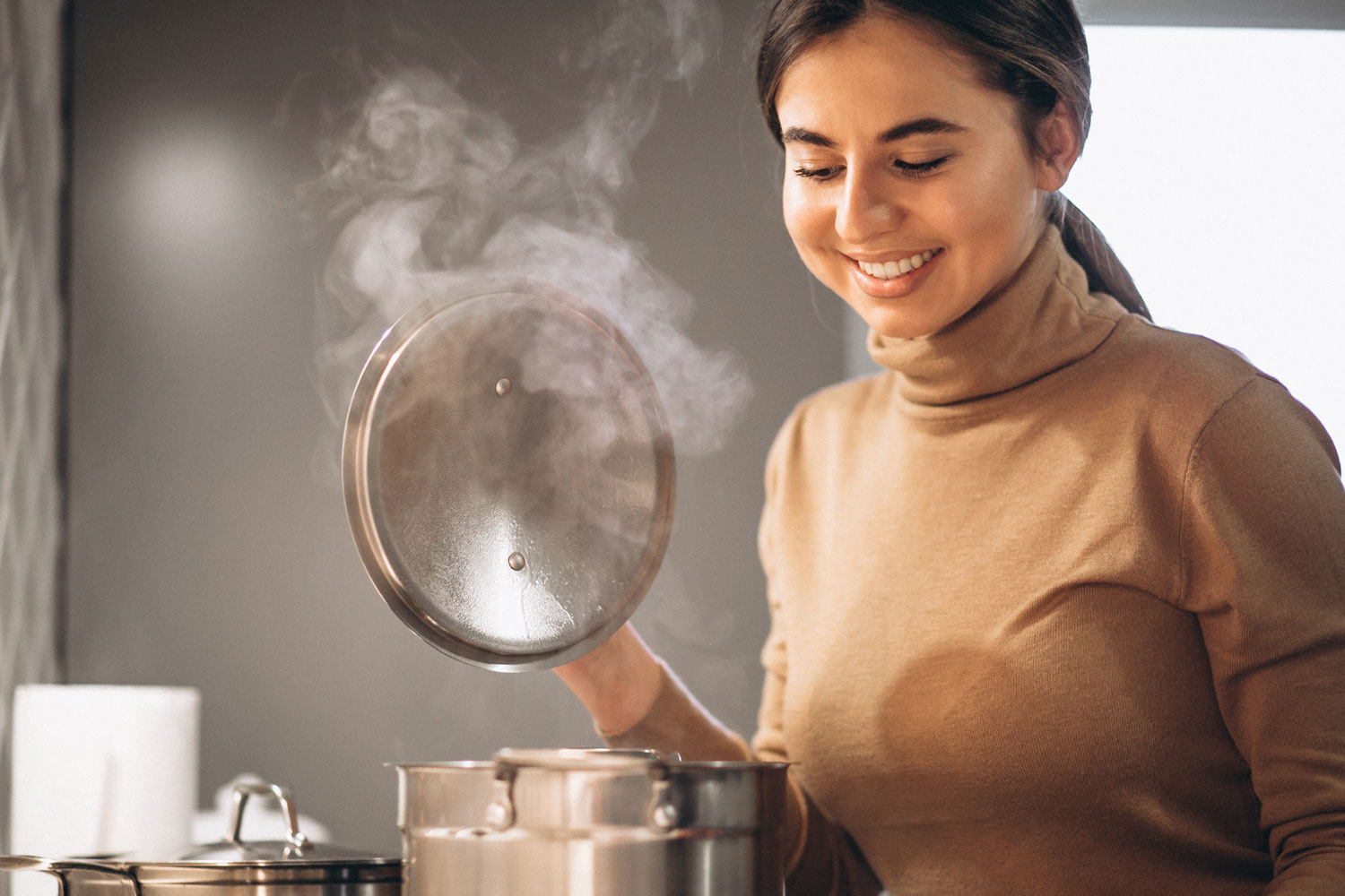 Seorang wanita memasak di dapur dengan uap keluar dari pancinya.