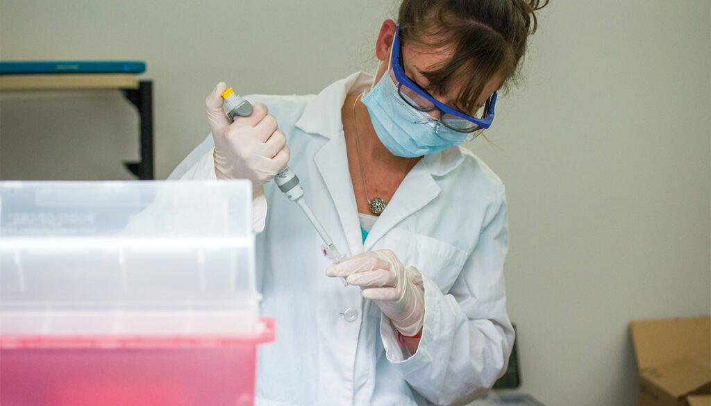 A person in a lab coat and mask holding a pipette

Description automatically generated