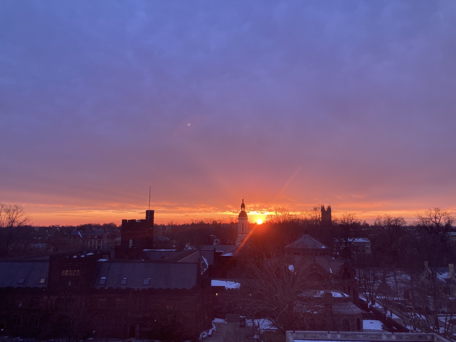 The sun sets creating a pink and purple sky behind a silhouette  of Nassau Hall and other campus buildings