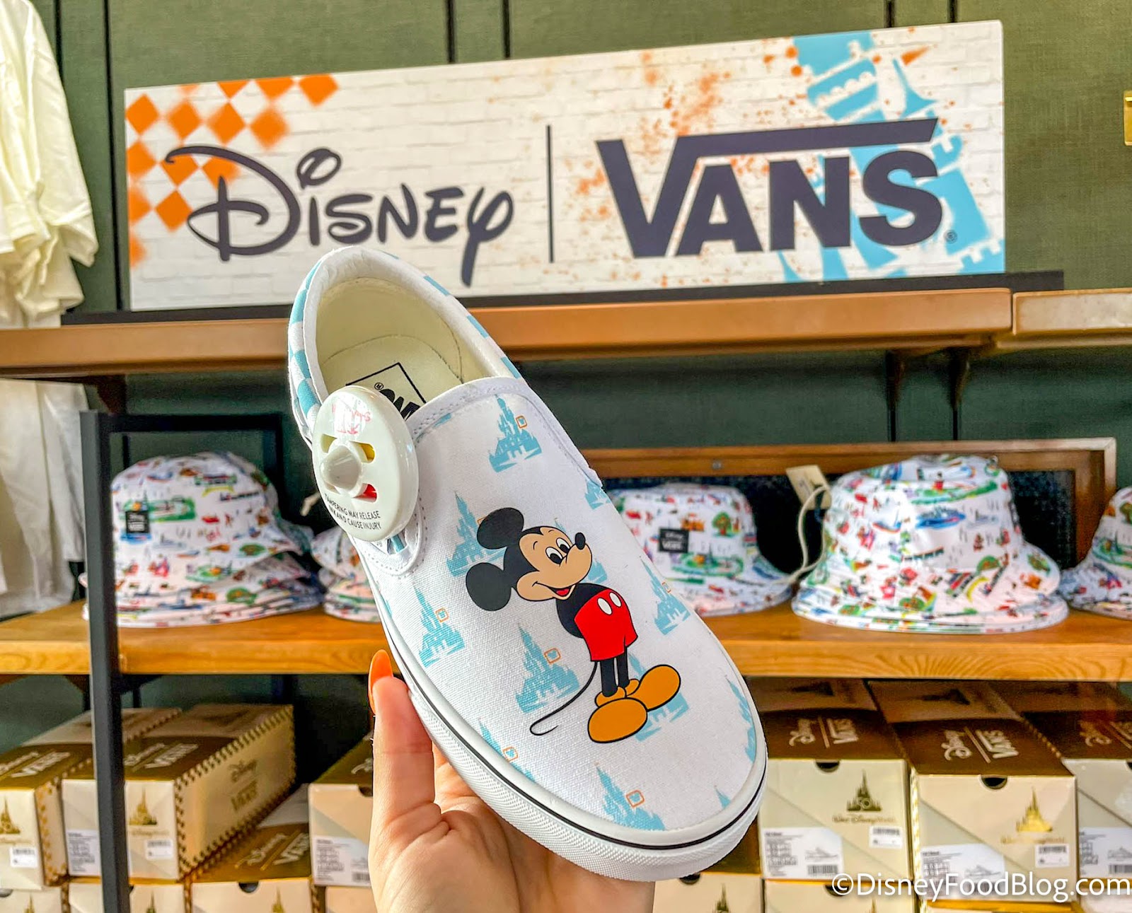Vans x Disney collaboration Mickey Mouse print slip-on shoe held in front of display with shoeboxes and Disney Vans signage in the background.